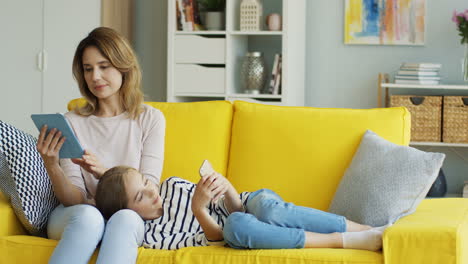 Caucasian-young-mother-and-teen-daughter-resting-on-the-sofa-at-home-and-using-the-tablet-device-and-smartphone.-Indoors.
